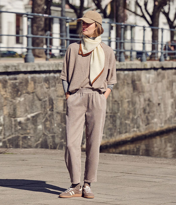 Woman wearing a beige corduroy co-ord with relaxed-fit trousers, matching top, cream scarf, baseball cap and sneakers.