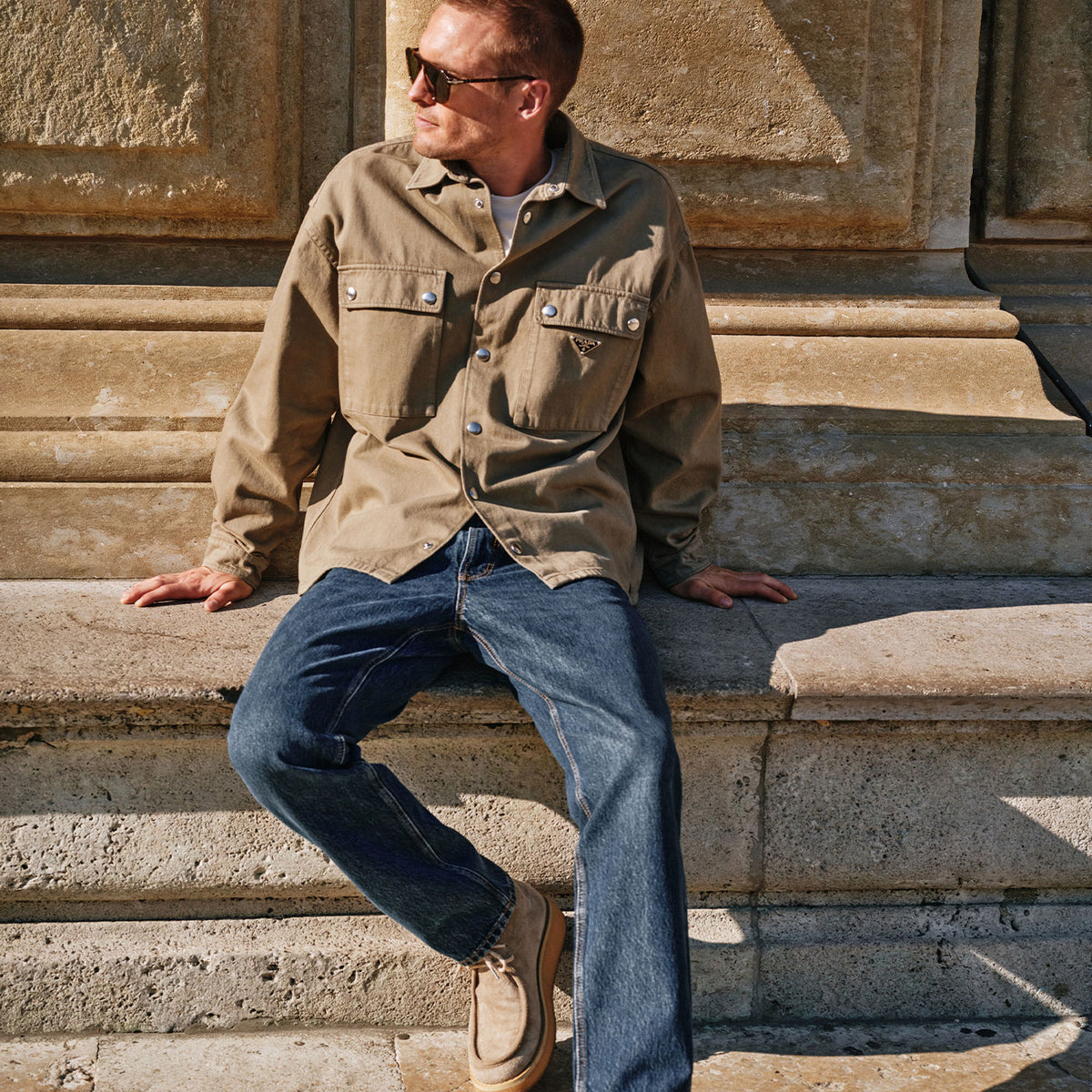 Man wearing dark blue Rick jeans paired with a beige jacket, seated casually on stone steps outdoors.