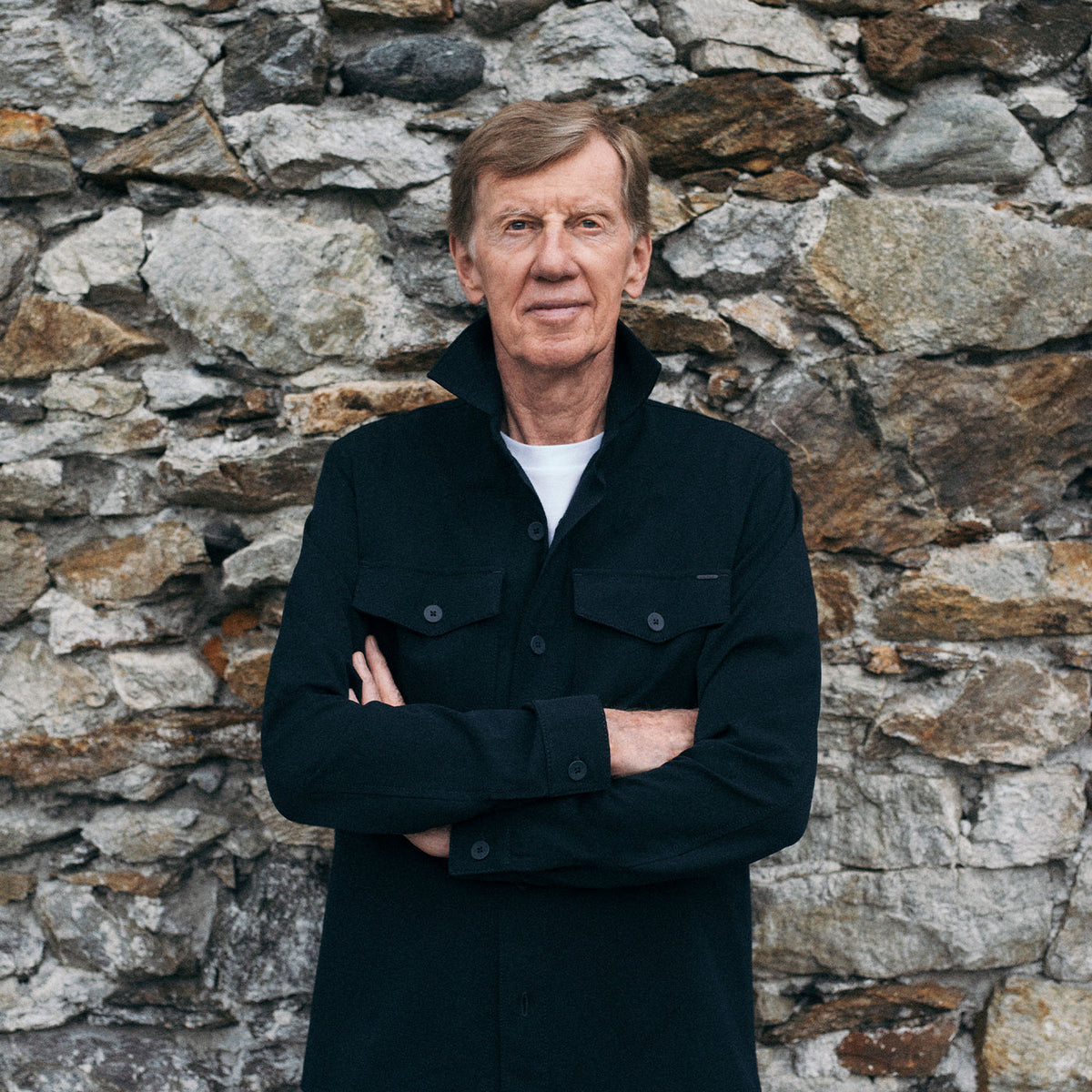 Walter Röhrl wearing a dark MAC Jeans jacket, standing with crossed arms in front of a stone wall.