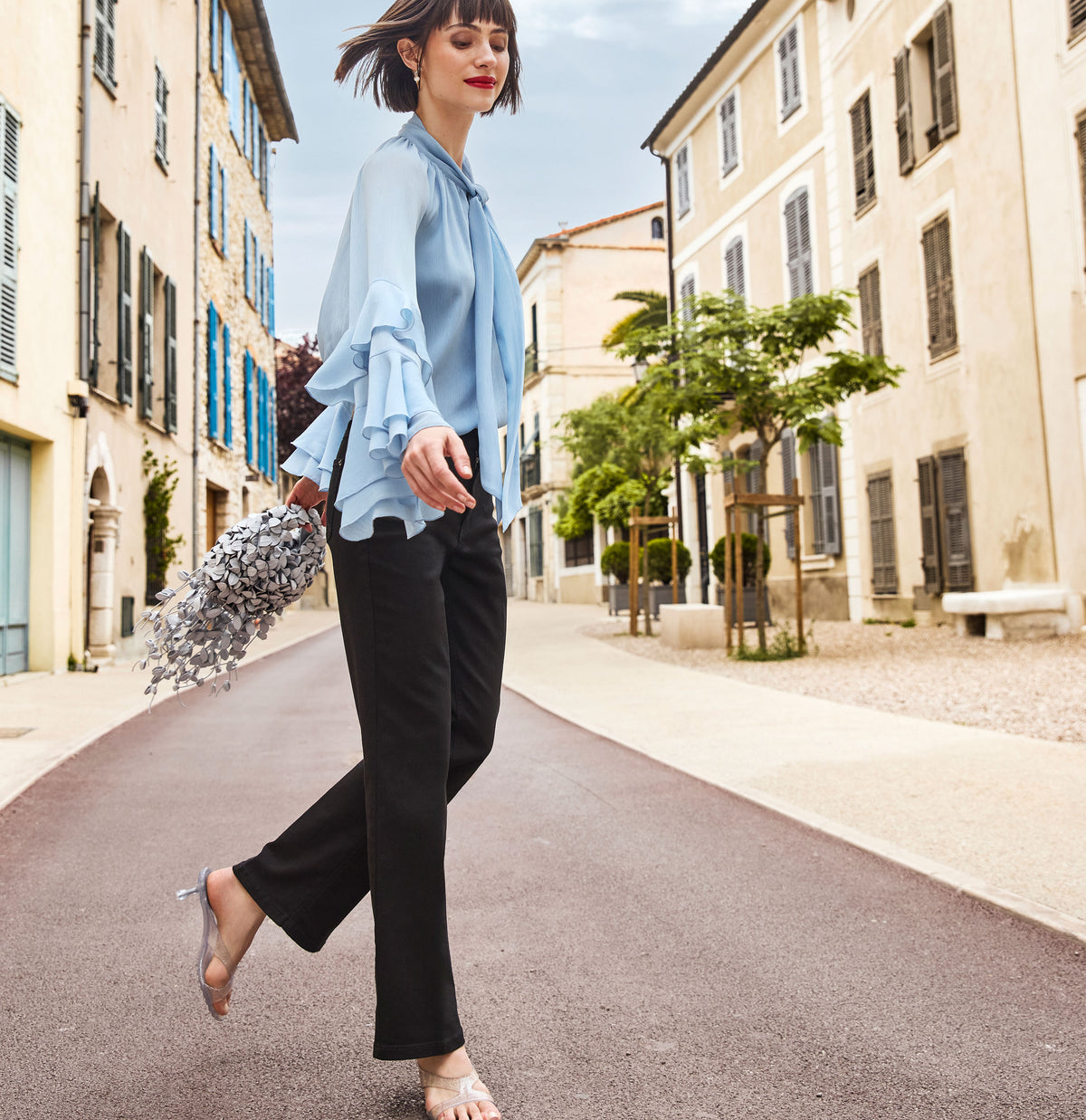 Model wearing Laura black trousers, a light blue blouse and metallic sandals walks through a Mediterranean old town street carrying a silver handbag.
