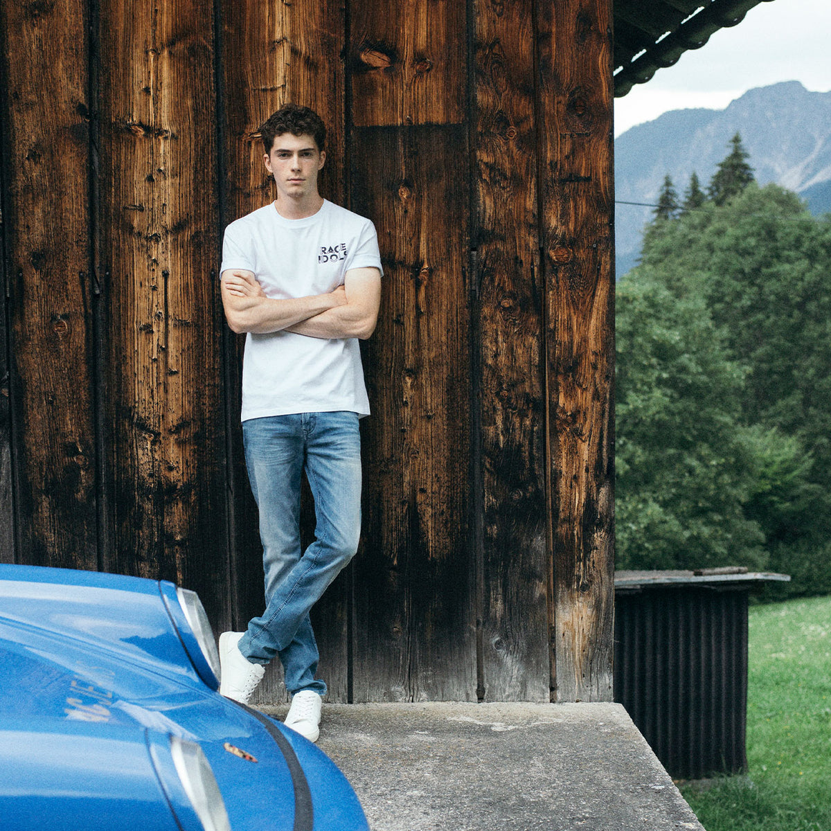 Valentin Kluss wearing MAC Driver Jeans and a Race Idols T-shirt beside a blue sports car in an alpine setting.