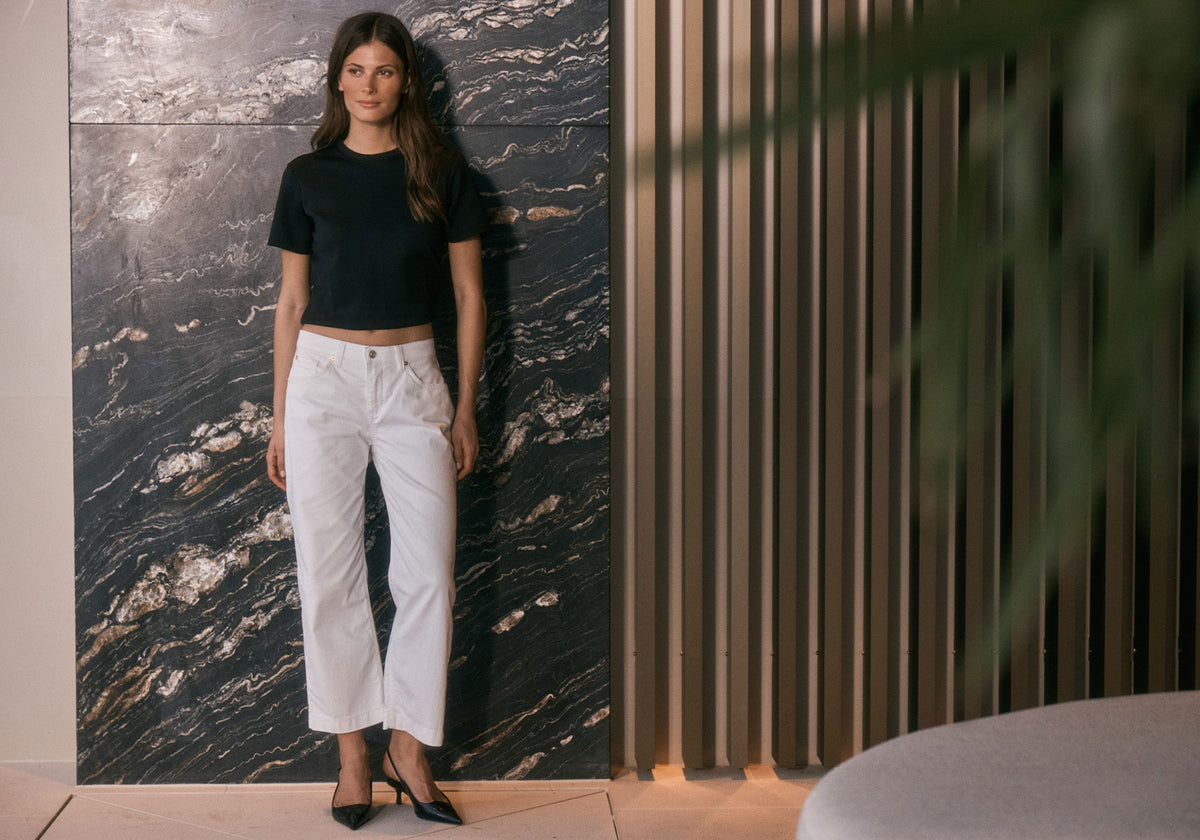 A woman in a black top and white cropped pants stands against a dark marble wall.