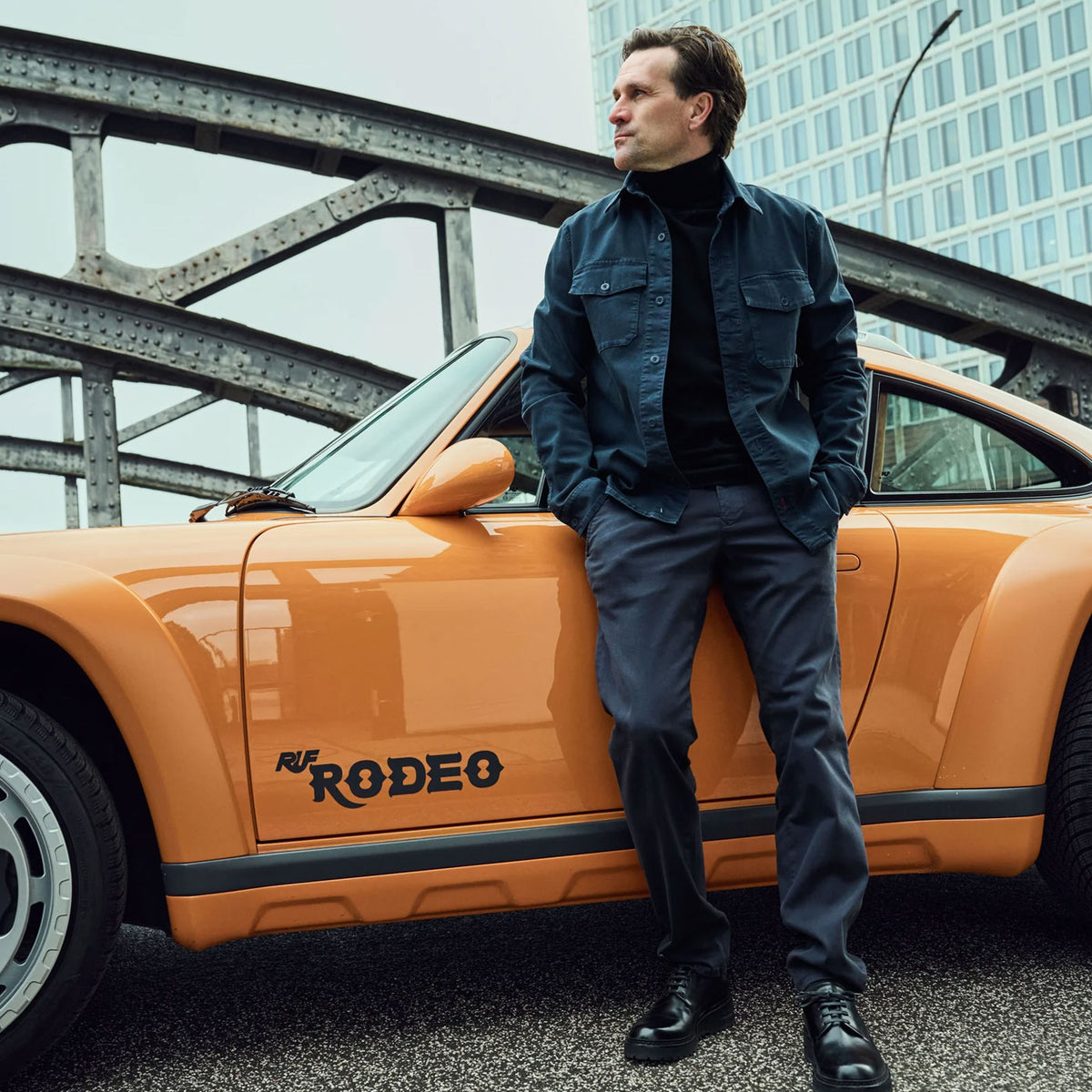 Markus Winkelhock wearing the MAC Ultimate Driver collection beside an orange classic sports car in an urban setting.