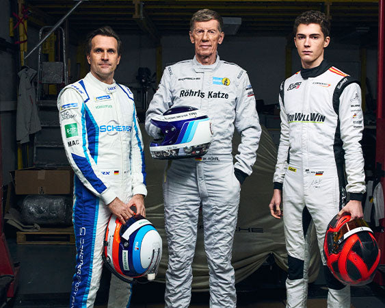 Three race drivers in white racing suits holding helmets, standing in a garage.