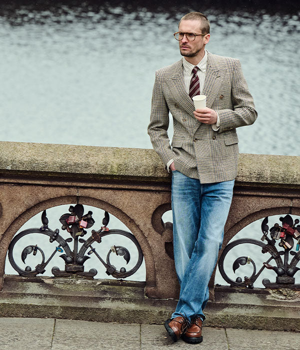 Man wearing light blue straight-leg jeans, a checked double-breasted blazer, white shirt, striped tie and brown leather shoes, leaning against a stone railing by the water.