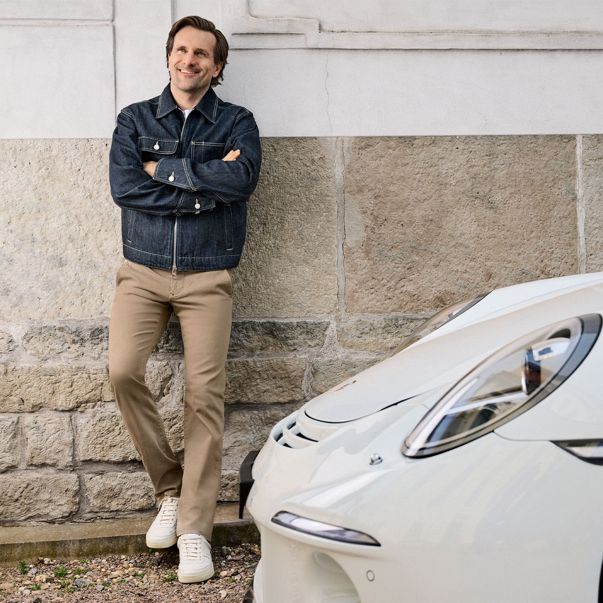 Man wearing a jacket and trousers, leaning against a wall outdoors.