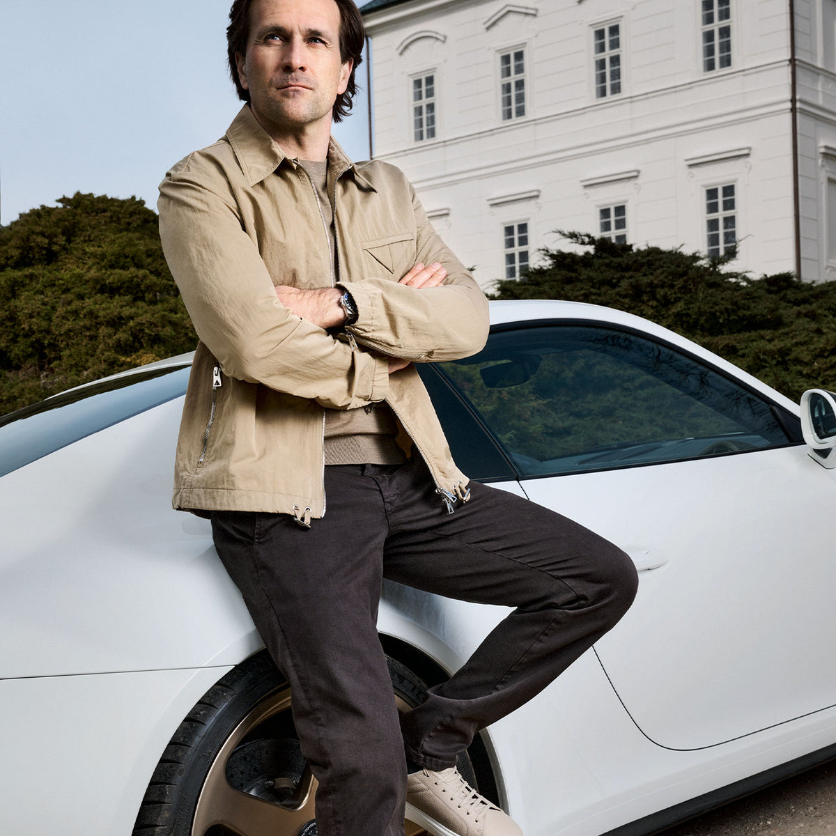Man wearing a jacket and trousers, leaning against a white sports car outdoors.